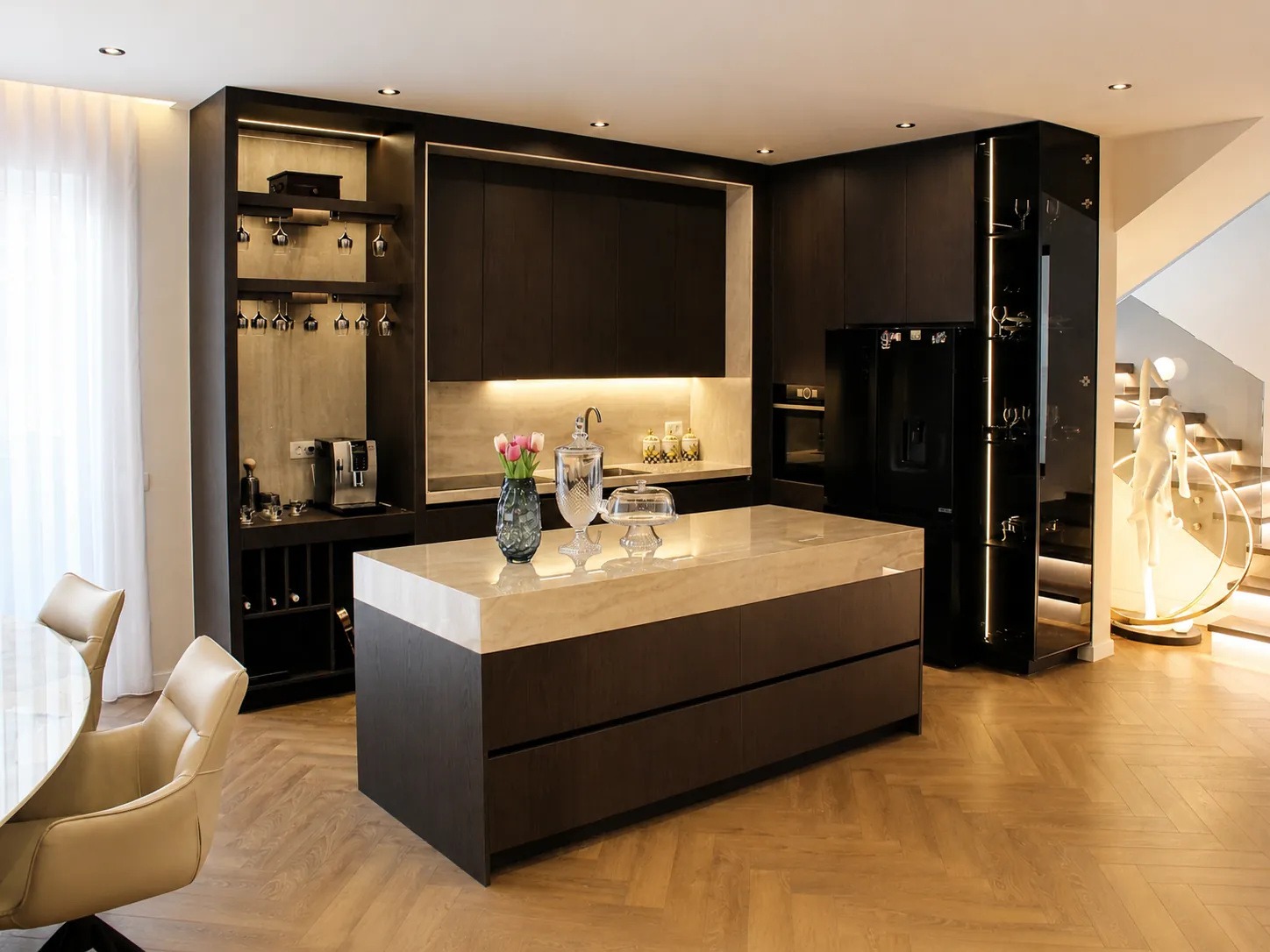 OR House as built: the kitchen wall with tall dark units, an island in stone and a lit glass cabinet.
